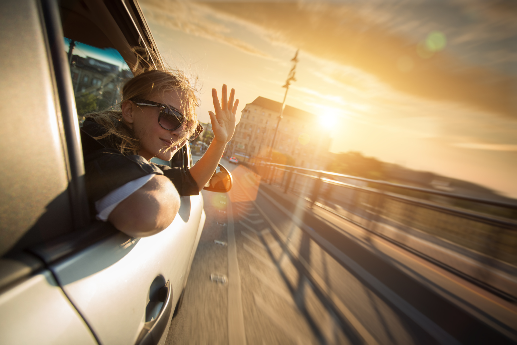 Junge Frau winkt aus geöffnetem Fenster eines fahrenden Autos.