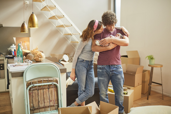 Ein Mann und eine Frau stehen in einer Wohnung zwischen offenen Kartons. Der Mann hält ein Baby im Arm.