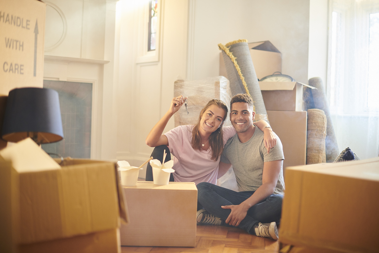 Zahnzusatzversicherung-umzug-ausland Paar sitzt Arm in Arm zwischen Umzugskartons. Sie hält einen Schlüssel hoch, beide lächeln und zeigen makellose Zähne.