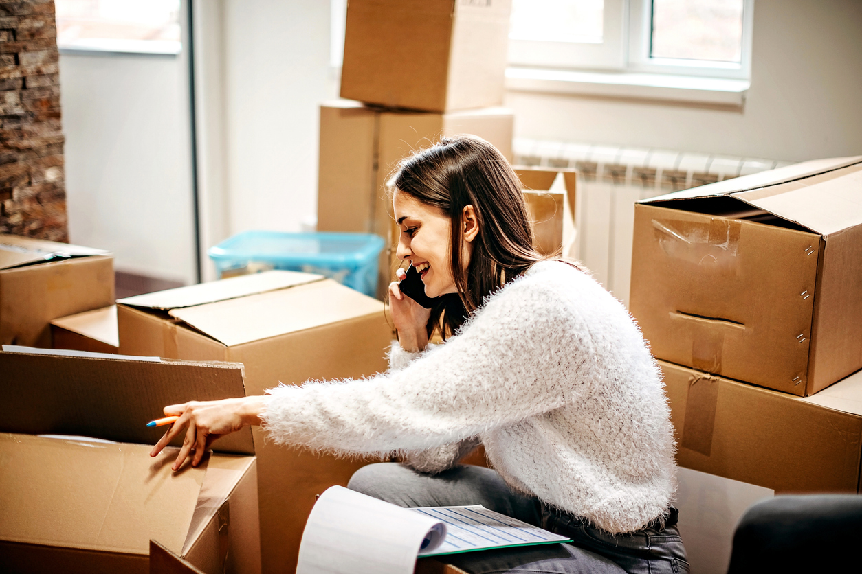 Berufsunfähigkeitsversicherung-Umzug-ins-Ausland Frau sitzt zwischen Umzugskartons und telefoniert. Sie beschriftet gerade einen Karton und lächelt.