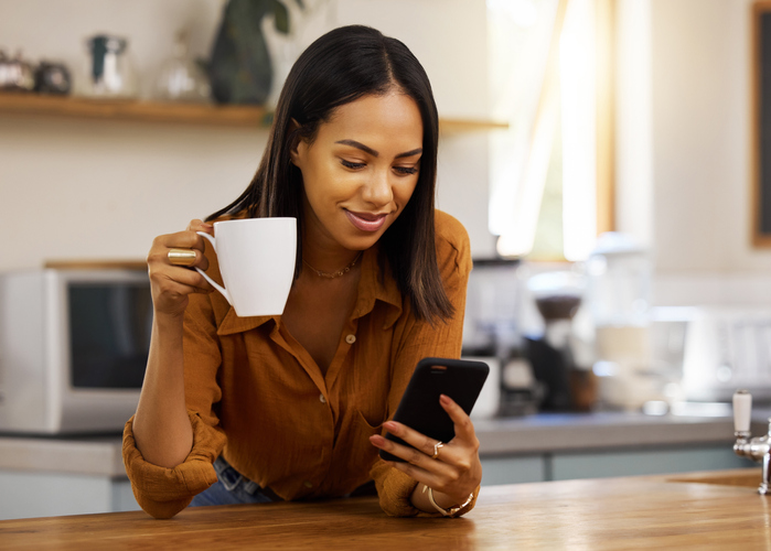 Wohnungssuche leicht gemacht: 6 Portale für die Suche im Internet Junge Frau hält eine weiße Tasse in der Hand, während Sie auf ihr Smartphone schaut.