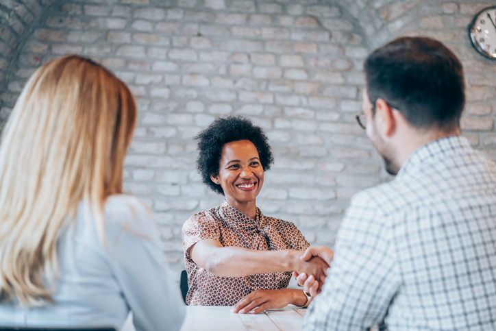 Mietkautionsbürgschaft: Was ist das und wie funktioniert sie? Eine lächelnde Frau schüttelt einem Mann über einen Tisch hinweg die Hand. Neben dem Mann sitzt eine weitere Frau mit langen Haaren.