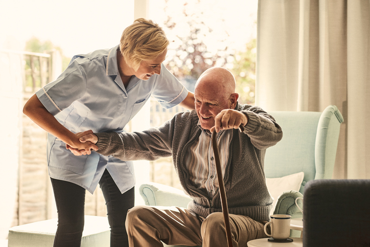 Senioren-WG: Modernes Lebenskonzept für Rentner und Best-Ager 50+ Eine Pflegerin hilft einem älteren Mann dabei, aus einem Sessel aufzustehen.