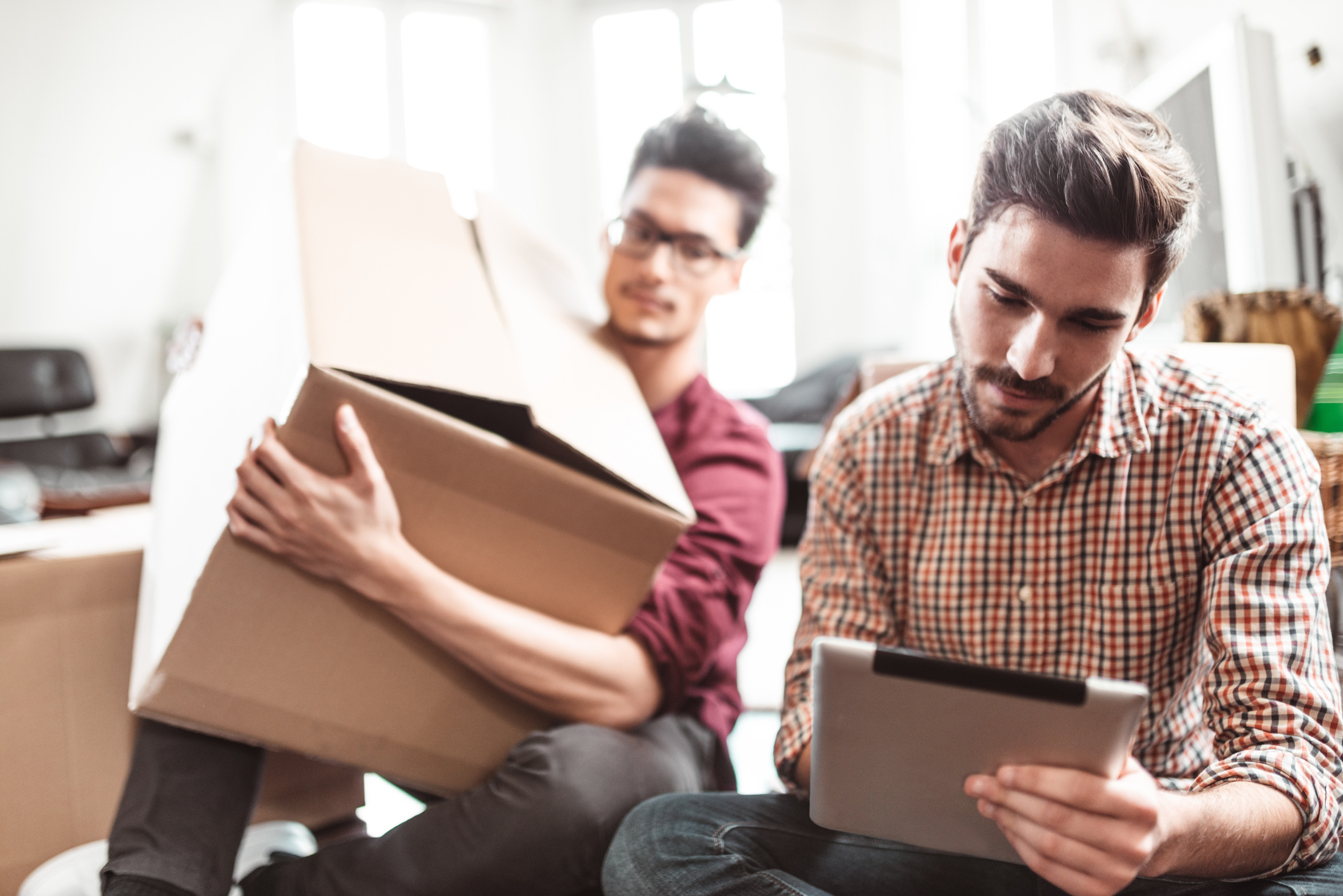 Mann blickt auf ein Tablet, im Hintergrund hält ein anderer Mann einen Karton in den Armen.