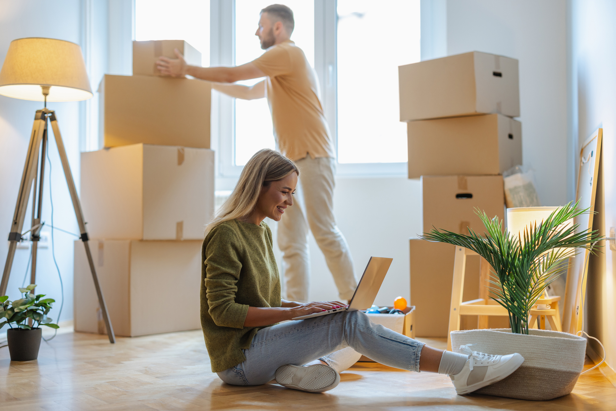 Frau und Mann in hellem Zimmer mit Umzugskartons und Lampe, sie blickt in Notebook. 