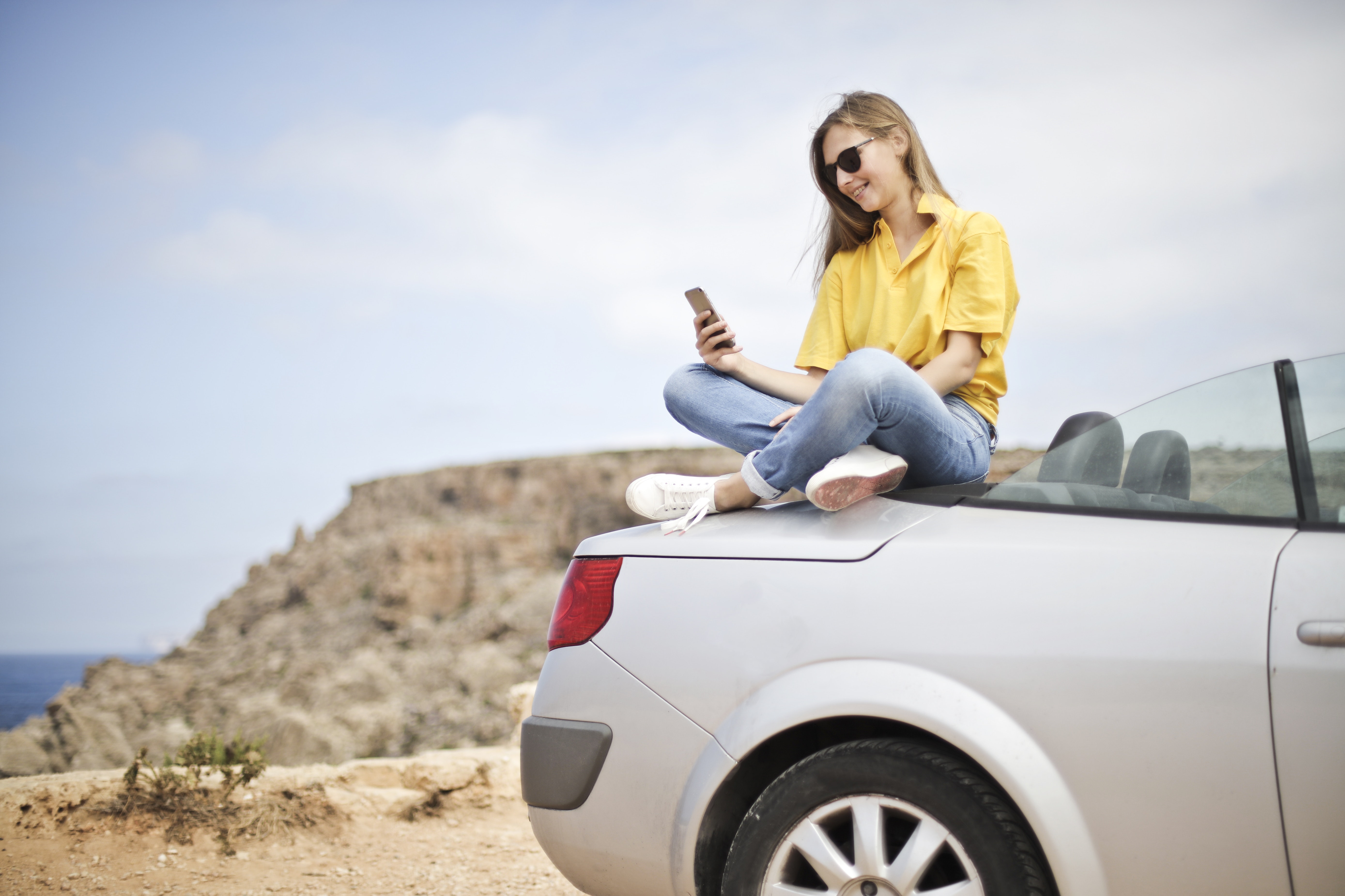 Auto online abmelden: So einfach geht's Junge Frau sitzt auf einem Auto und schaut aufs Handy
