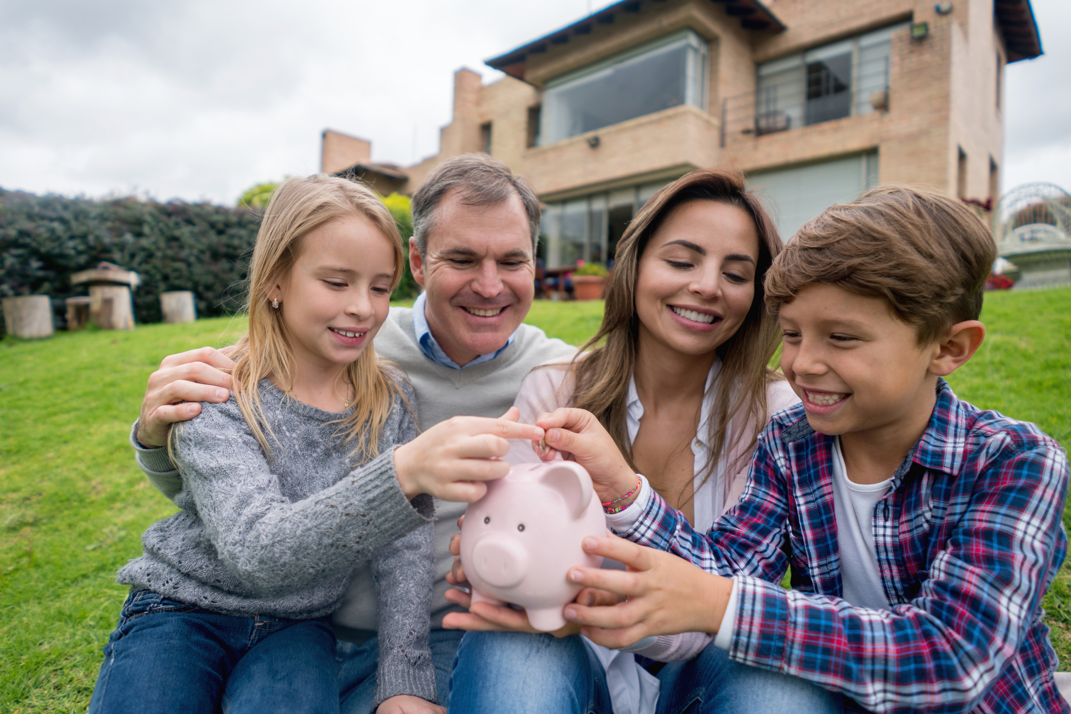Eltern mit zwei Kindern sitzen vor ihrem Haus und werfen gemeinsam eine Münze in ein Sparschwein
