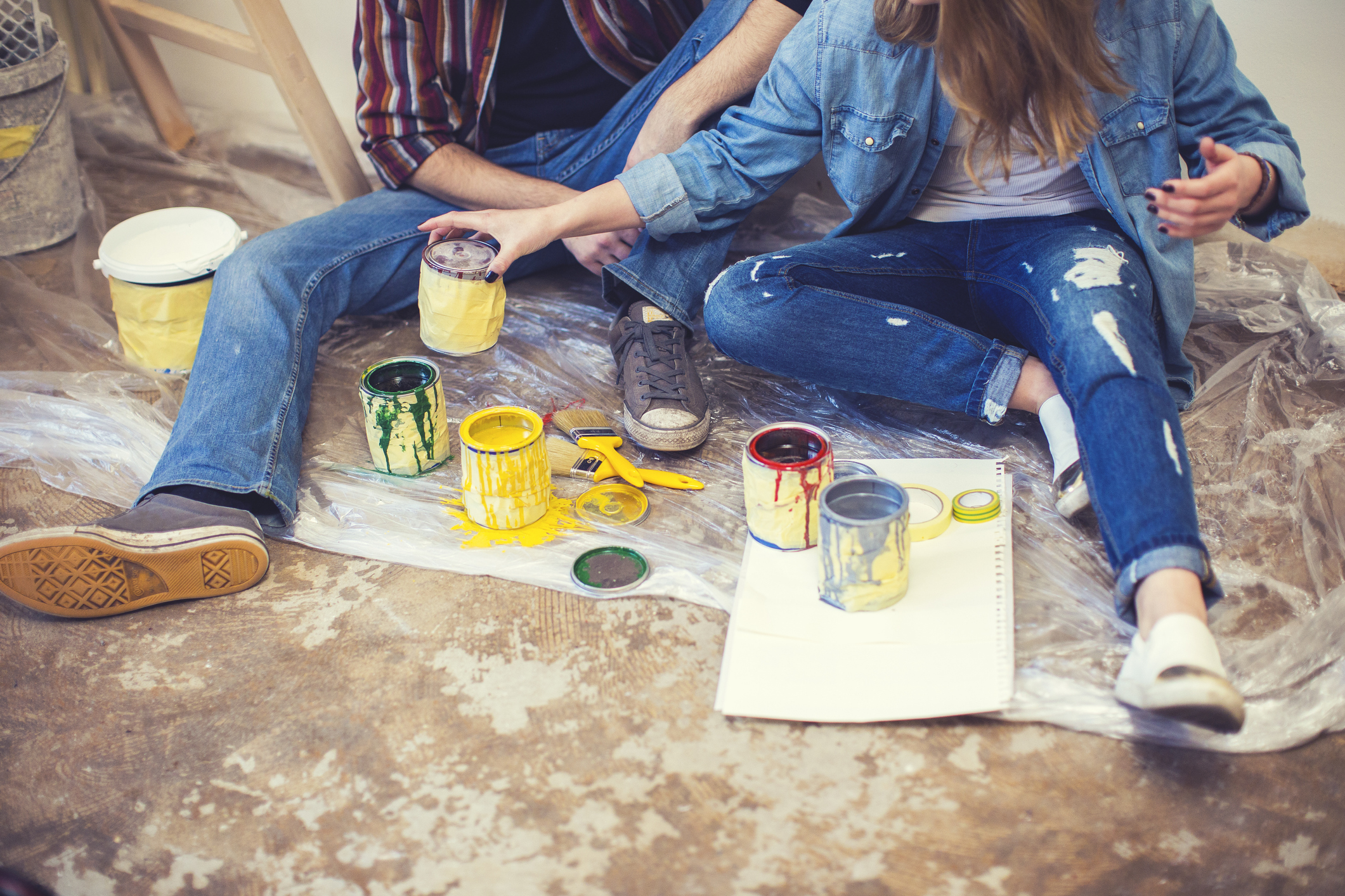 Farbe und Farbeimer entsorgen: Wohin damit nach der Renovierung? Junges Paar sitzt auf dem Boden zwischen leeren Farbeimern und Farbdosen