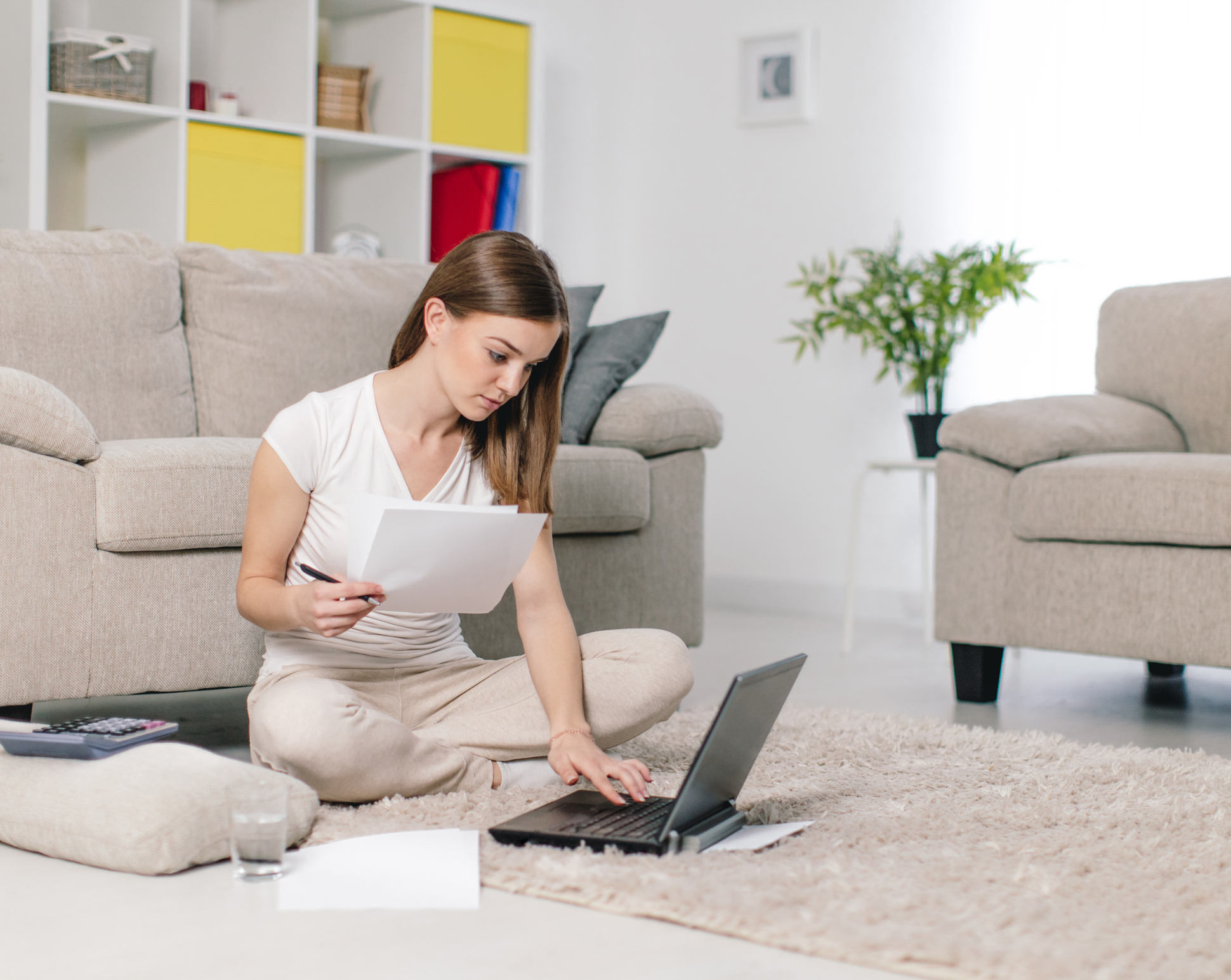 Junge Frau sitzt mit Laptop und Formularen auf dem Boden im Wohnzimmer