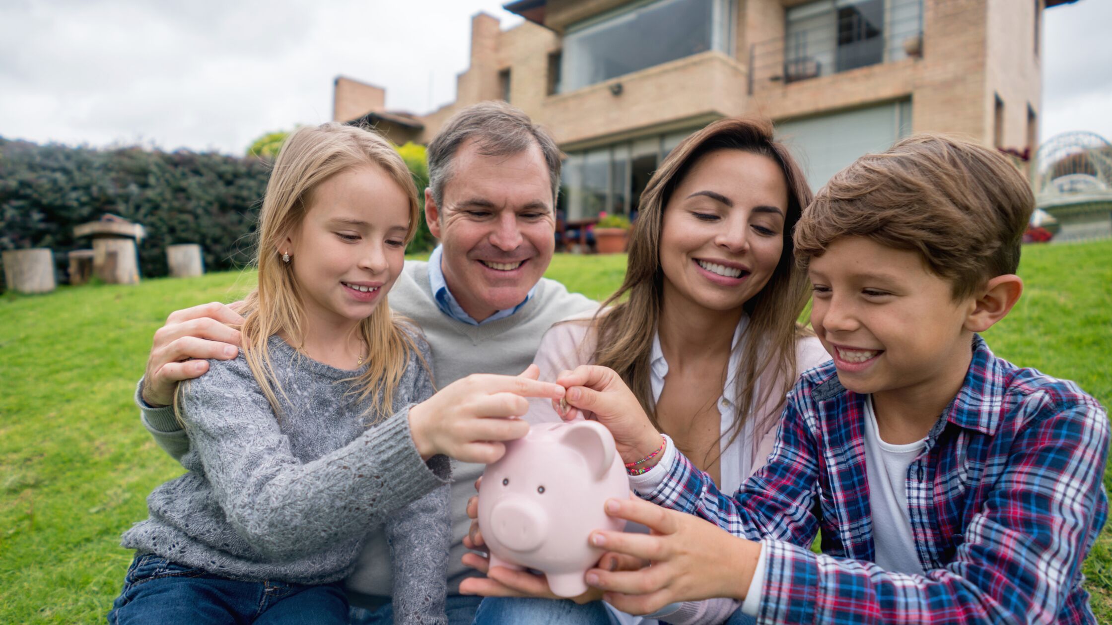 Eltern mit zwei Kindern sitzen vor ihrem Haus und werfen gemeinsam eine Münze in ein Sparschwein