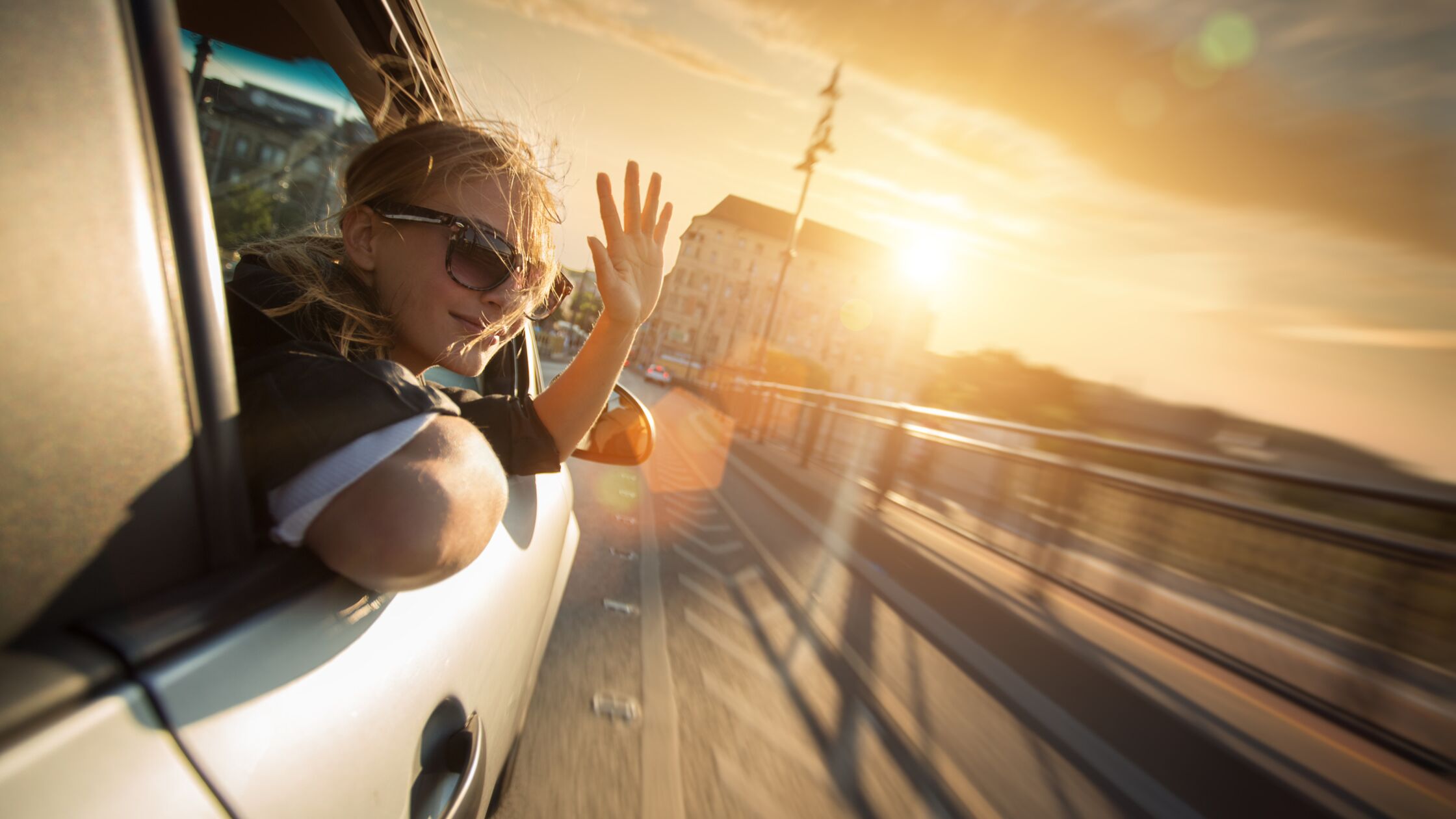 Junge Frau winkt aus geöffnetem Fenster eines fahrenden Autos.