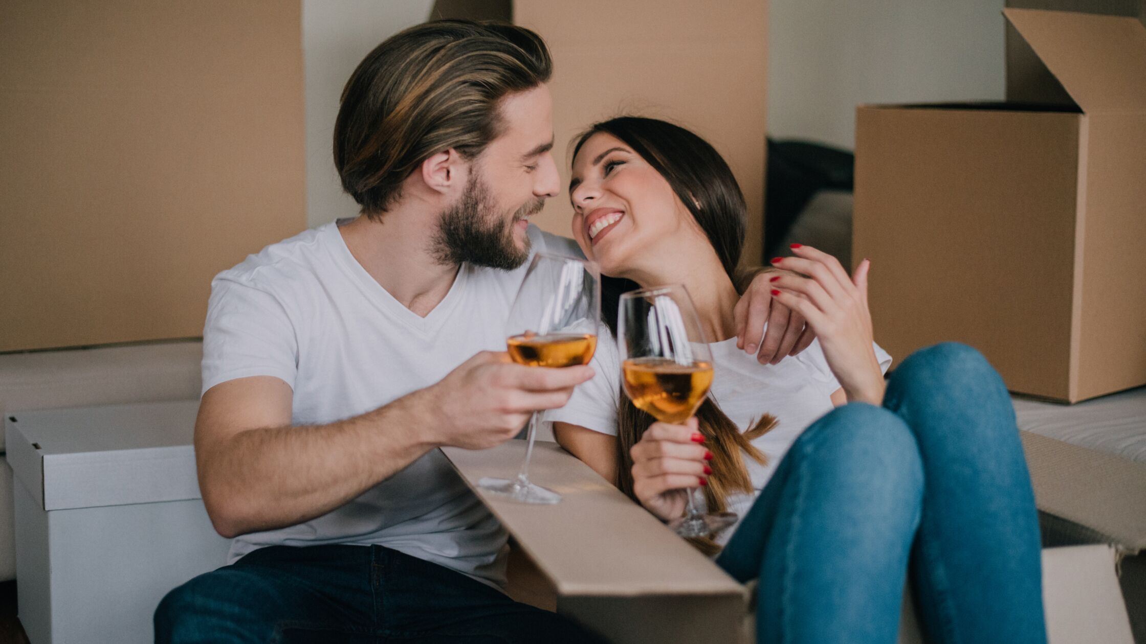 Im Hintergrund Umzugskisten, ein Paar stößt lächelnd mit Wein an.