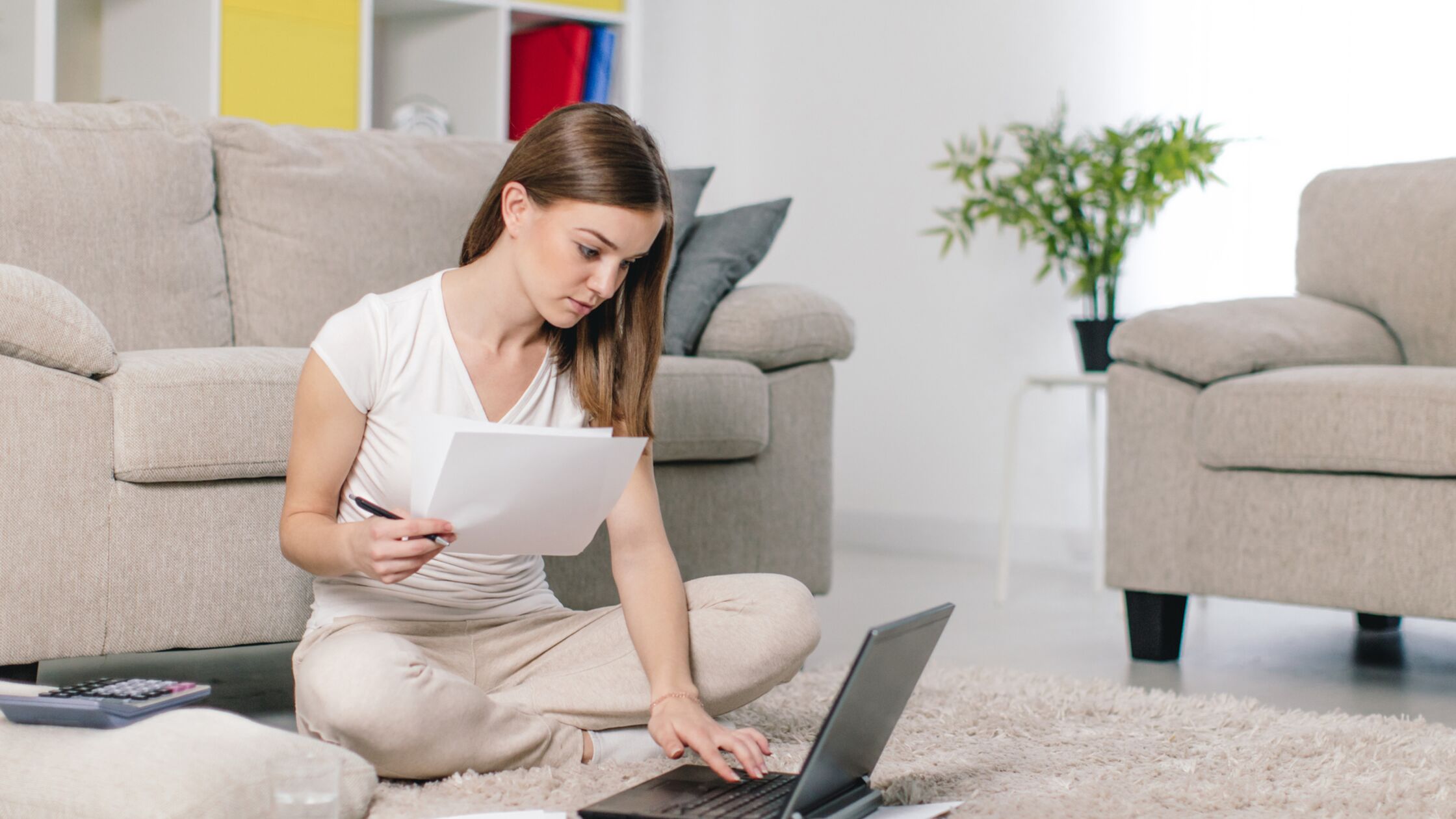 Junge Frau sitzt mit Laptop und Formularen auf dem Boden im Wohnzimmer