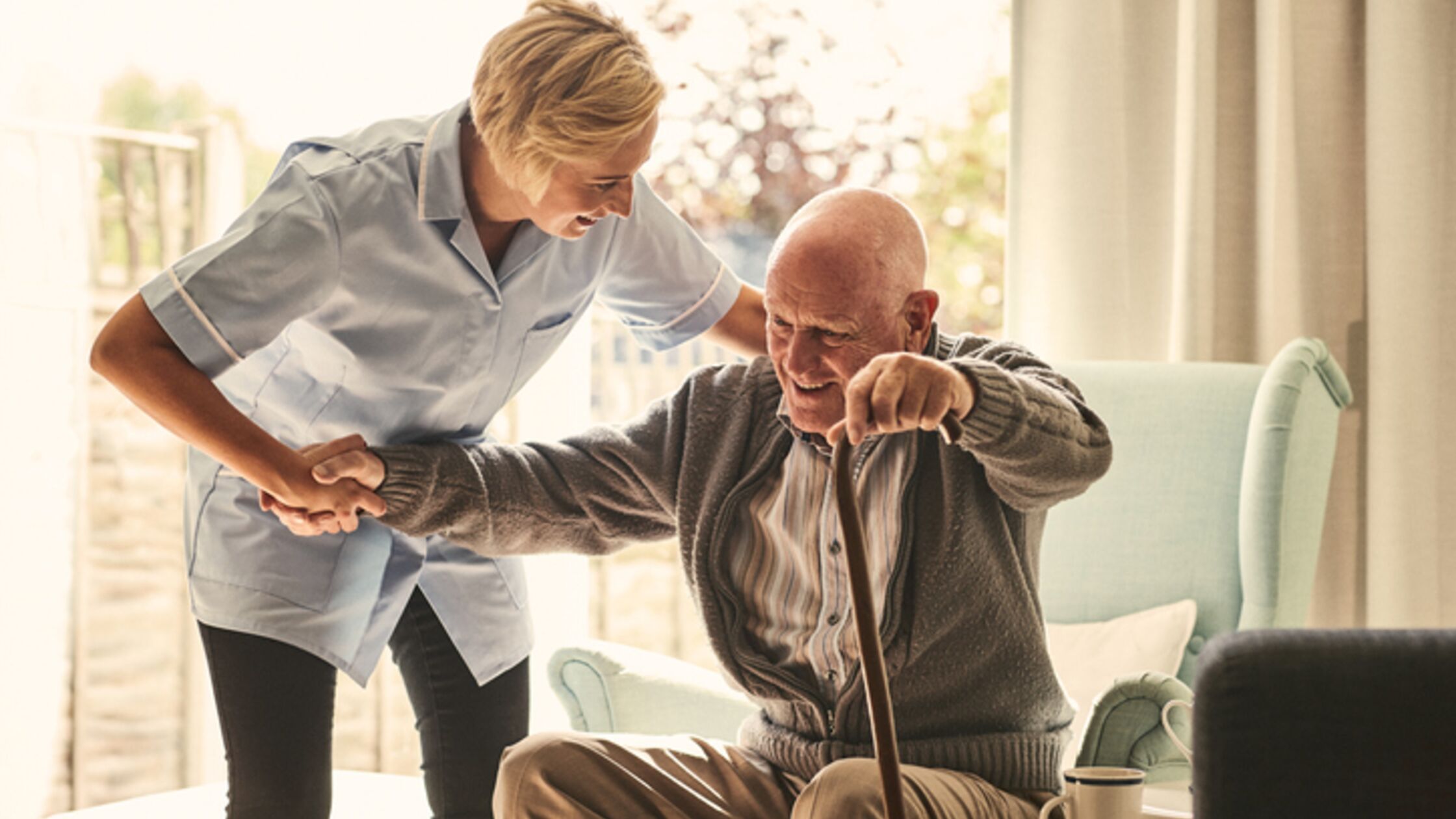 Eine Pflegerin hilft einem älteren Mann dabei, aus einem Sessel aufzustehen.