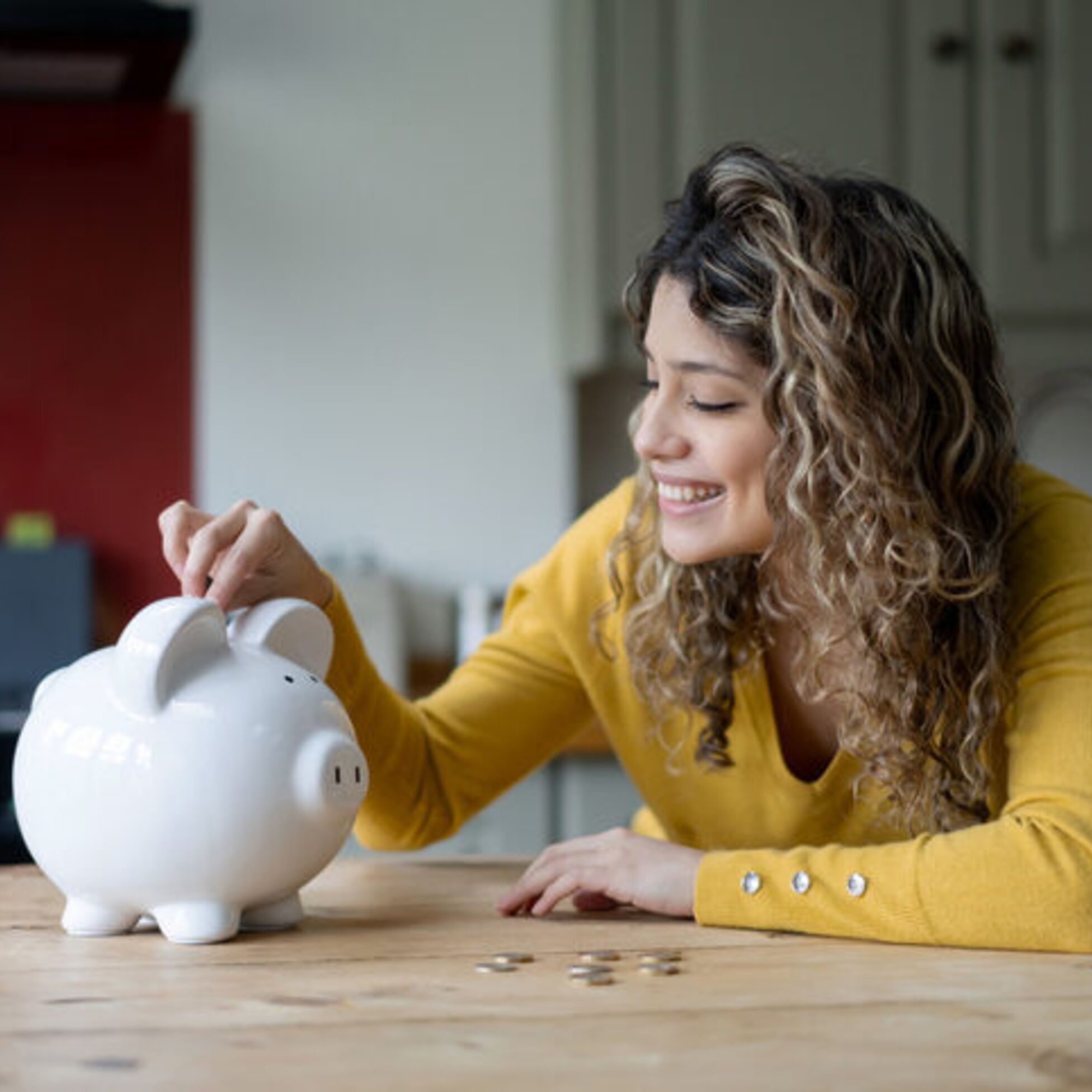 Junge Frau sitzt am Tisch und steckt lächelnd Münzen in ein weißes Sparschwein