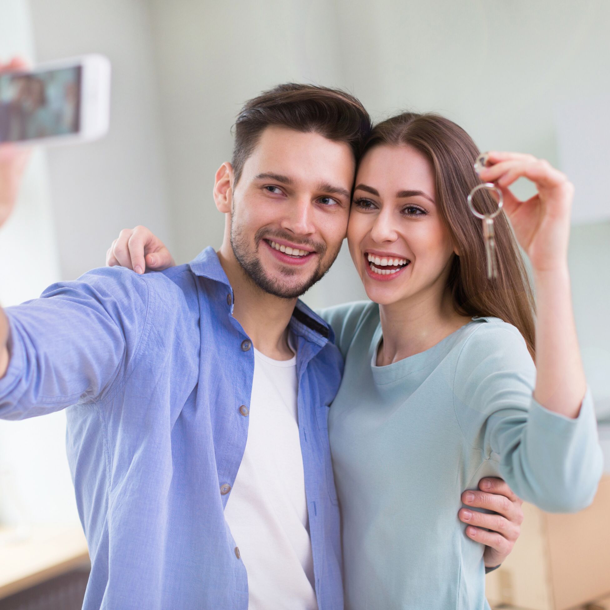 Junges Paar hält Schlüssel zu neuer Wohnung hoch und macht Selfie