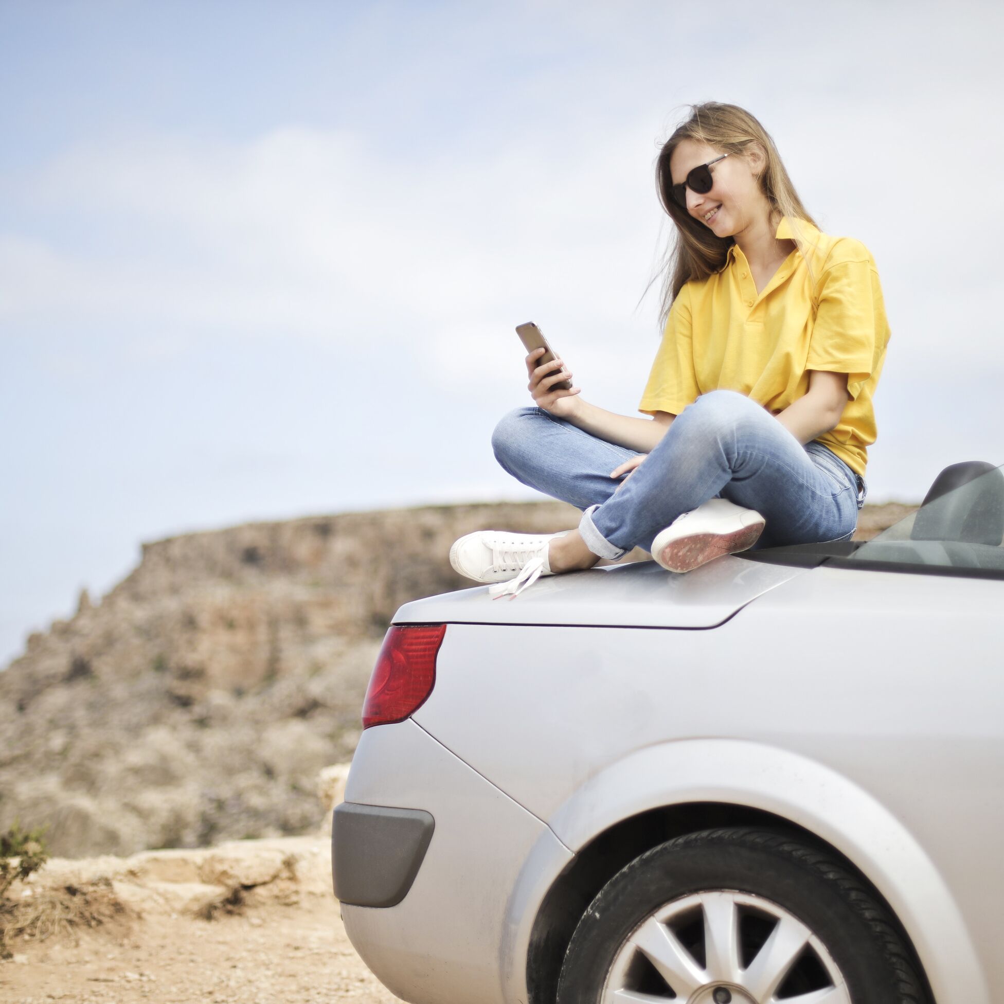 Junge Frau sitzt auf einem Auto und schaut aufs Handy