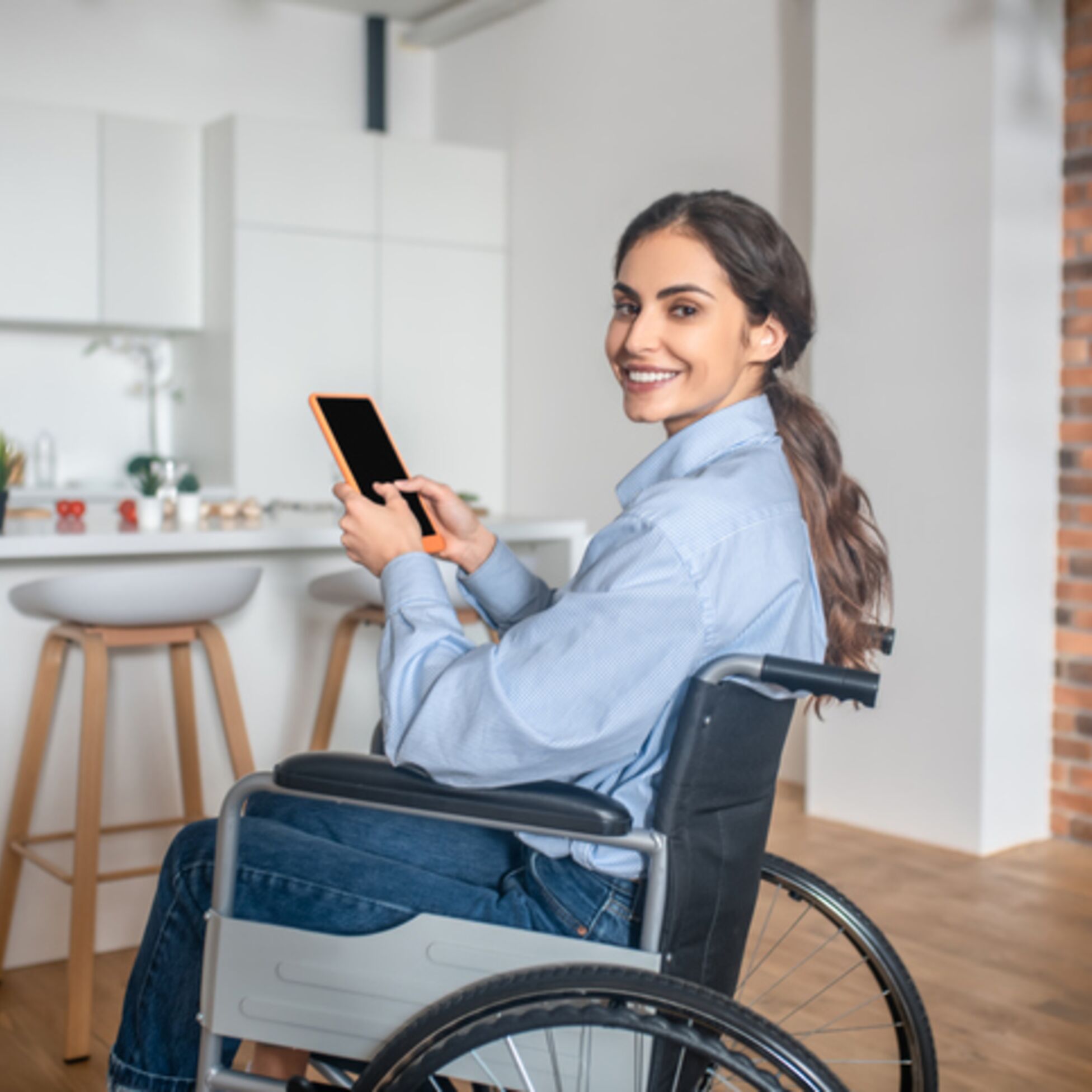 Junge Frau sitzt im Rollstuhl und hält ein Tablet in der Hand, im Hintergrund ist eine Küche